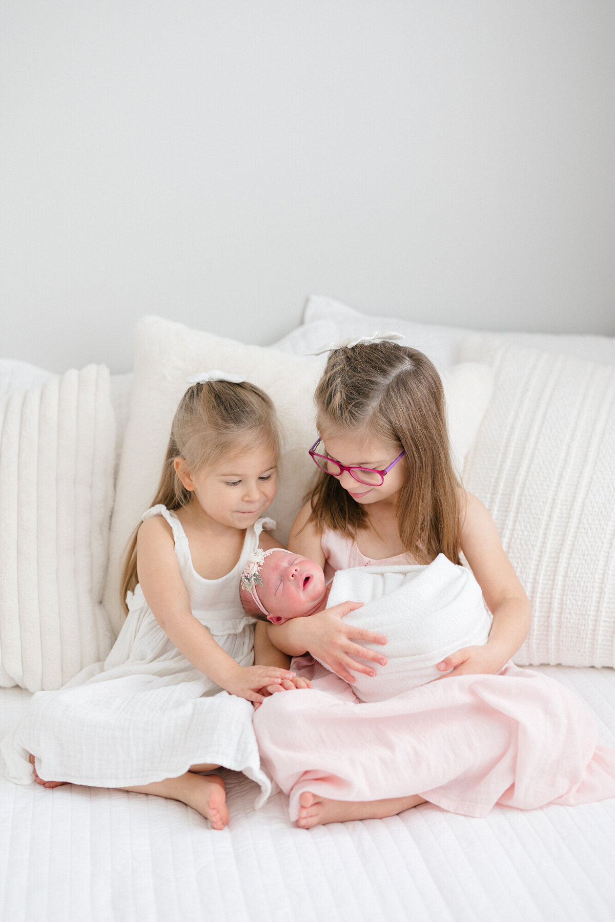 Two sisters dressed in pink and white dresses holding their newborn baby sister taken by louisville newborn photographer missy marshall