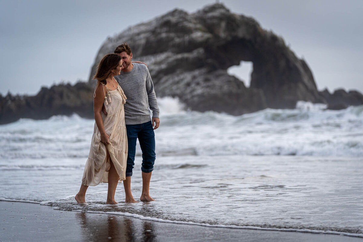 couple wading through the surf on the beach with a rock in the background containing a heart shaped cutout