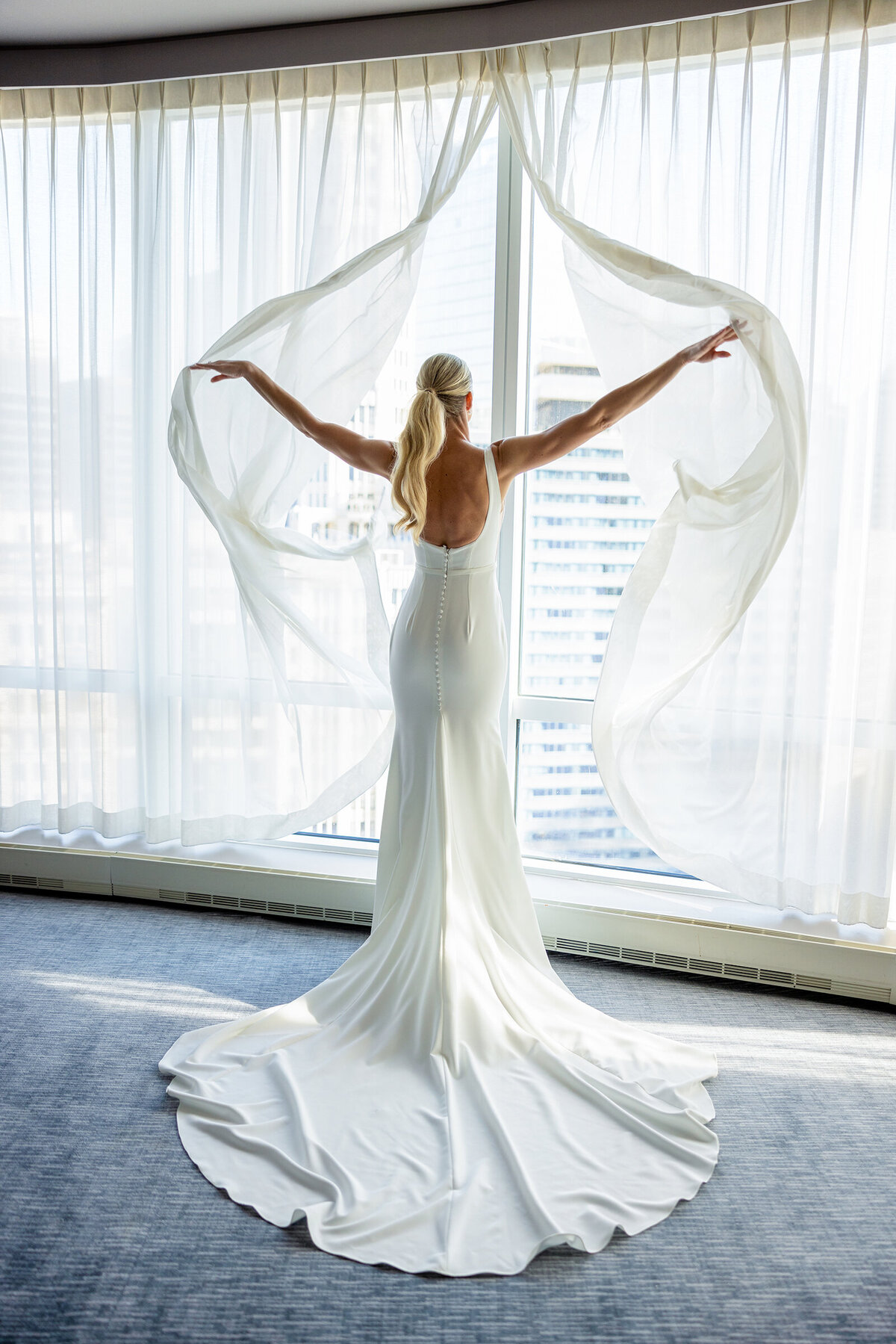 Elegant bride dressed in Made with Love designer wedding dress opening white window curtains at the trump hotel in Chicago