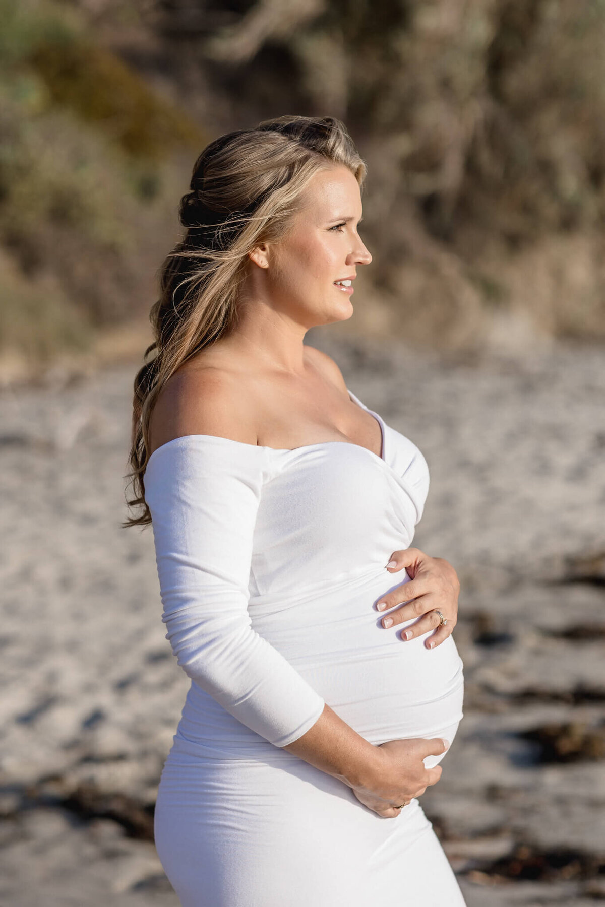 A pregnant woman in a white off-shoulder dress stands on a sandy beach, gently holding her belly and gazing into the distance. Her long hair flows in the breeze, and the background features blurred green foliage and sand.