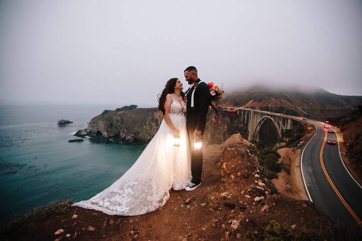 The bride and groom hold lanterns with Bixby Bridge in the background