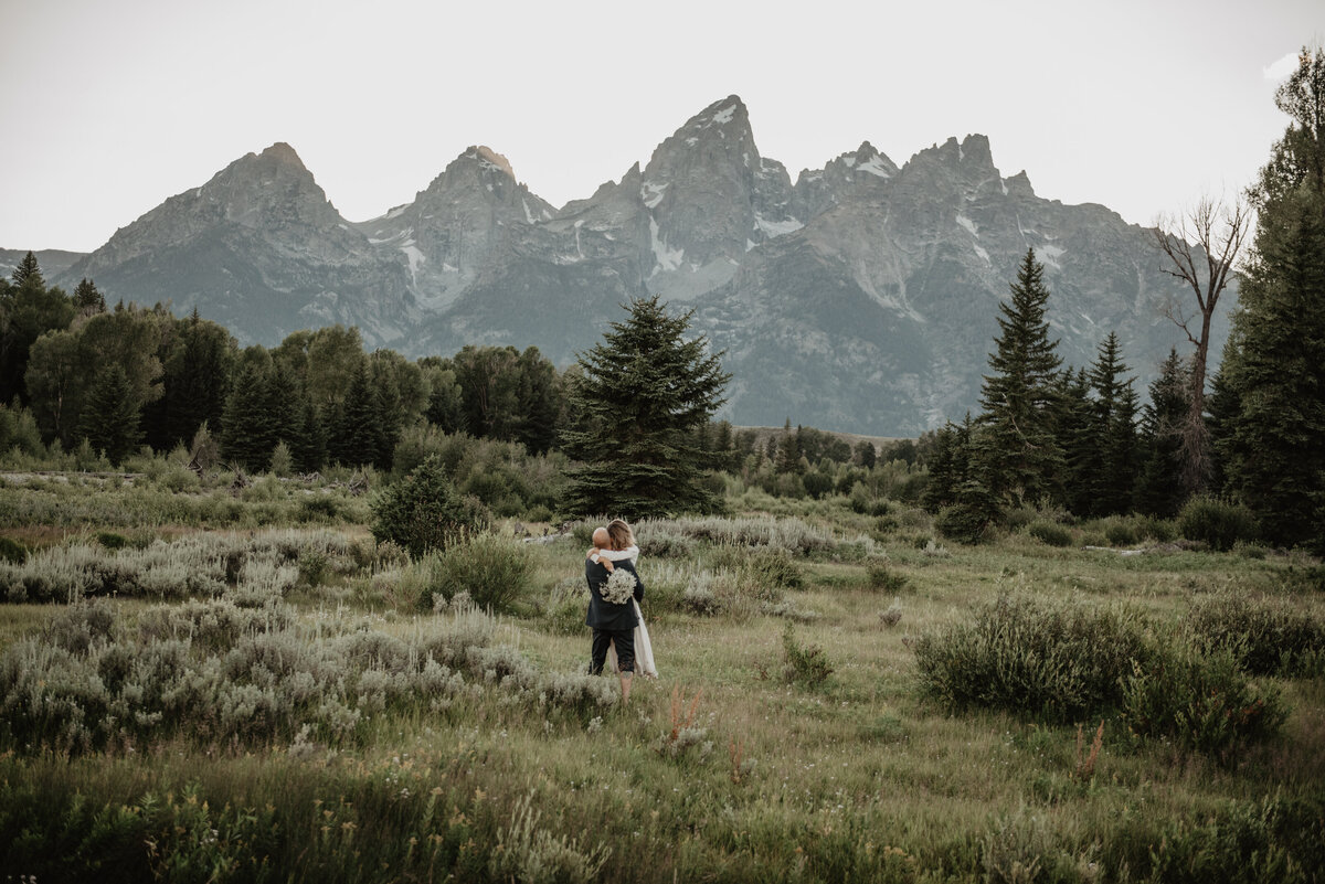 Grand Teton wedding day with man carrying bride through a sage bush field in front of Tetons