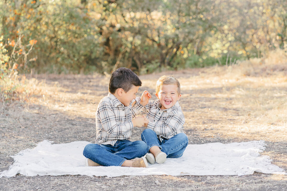 two young brothers sit on a white blanket at an Austin park and laugh together.