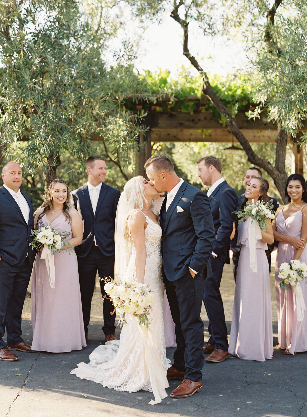 A romantic photo of a bride and groom sharing a kiss surrounded by their bridal party at Holman Ranch in Carmel Valley, California. The wedding party is dressed in coordinated attire, celebrating a beautiful outdoor wedding at this stunning venue