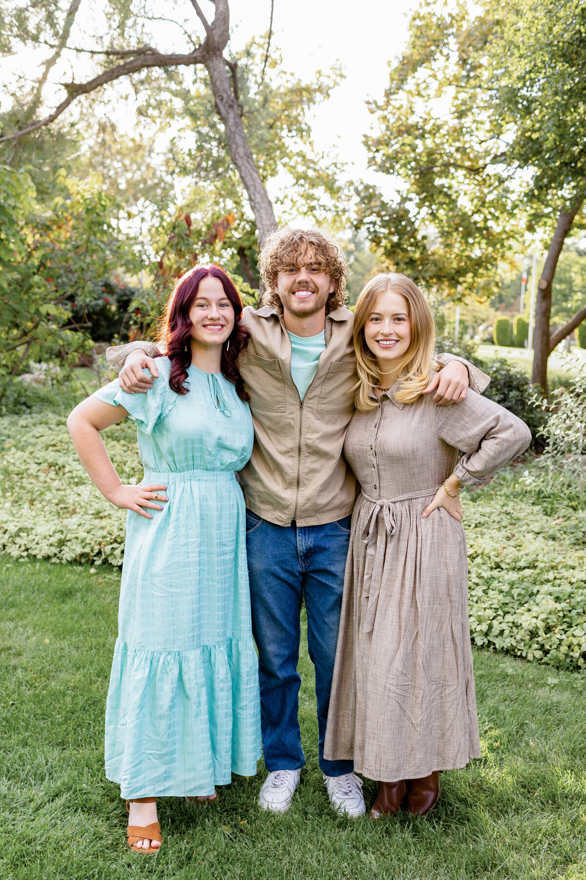 Three siblings stand close together, arms around each other, smiling against a backdrop of trees and shrubs.