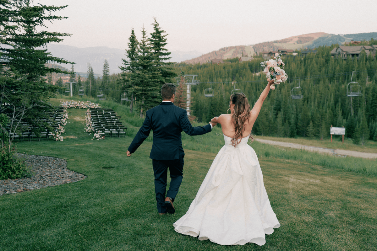 Bride and groom walking hand in hand at their Moonlight Basin wedding, with the bride raising her bouquet and a floral-lined mountain ceremony site in view.