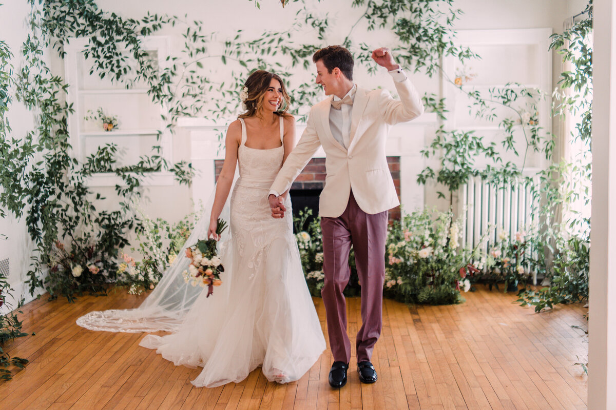 Bride wearing Alexandra Grecco & groom in white & rose tuxedo cheering during recessional in room filled with vines & flowers