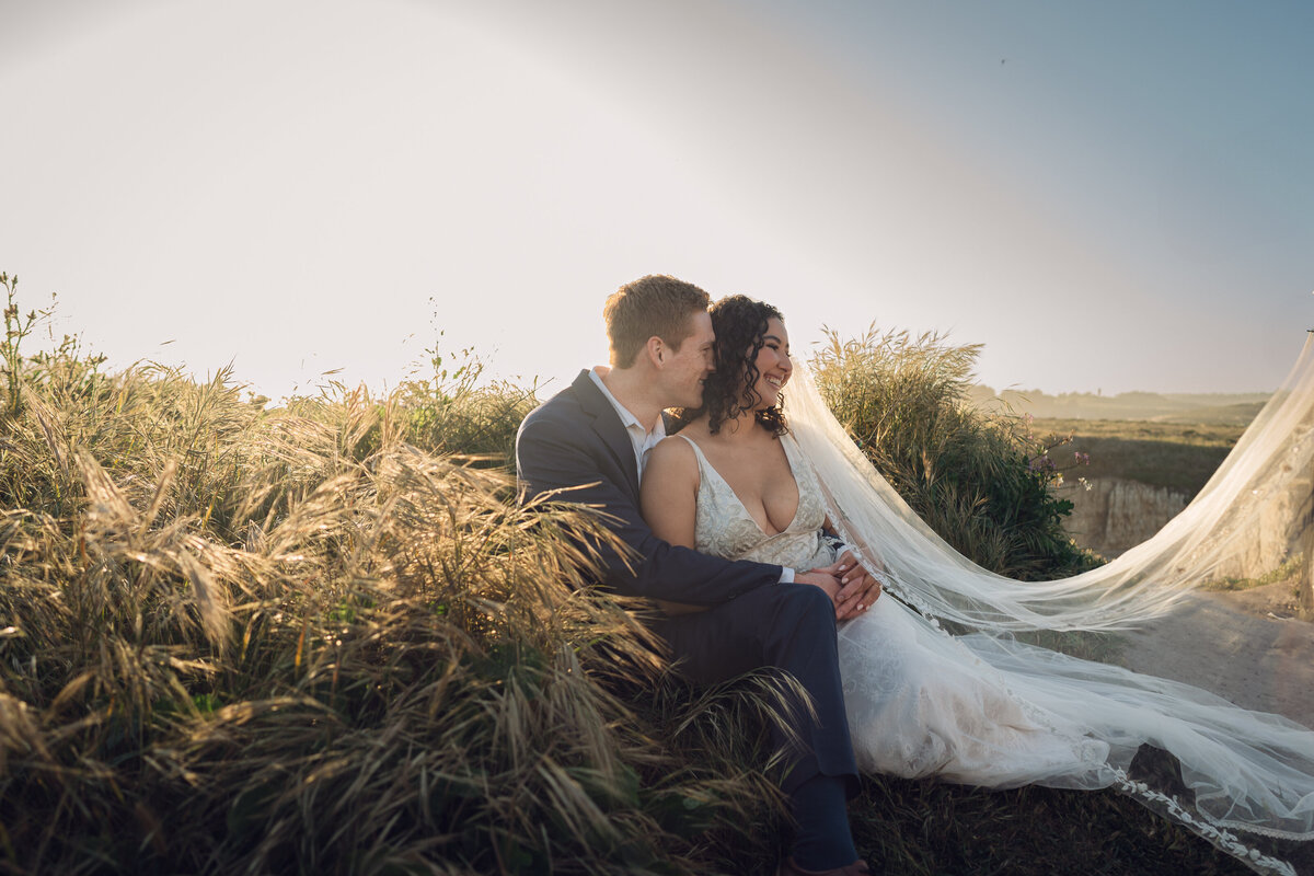Bride and groom on meadow in Santa Cruz at golden hour sitting in the long grass with the bride's veil cascading over her