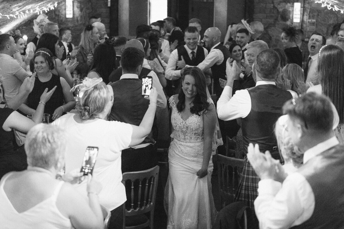 A bride and groom walk through a room of guests to take their place at the top table. The guests cheer their arrival.
