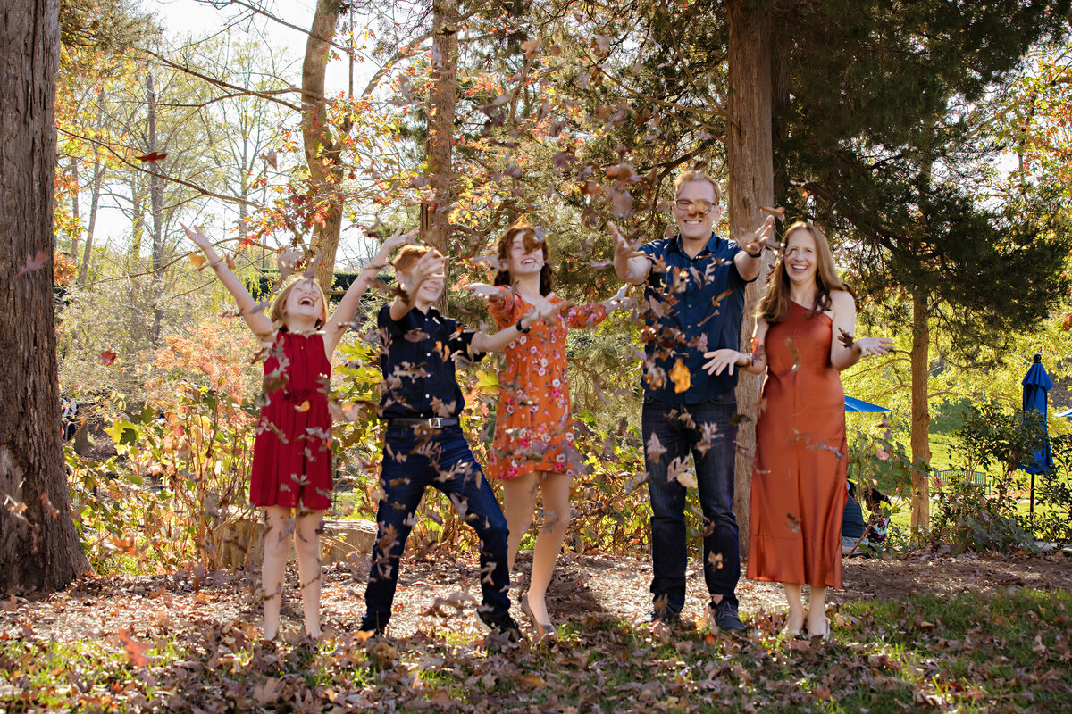 Vivacious family moment with parents and three kids joyfully throwing piles of golden leaves in a wooded park at Ballantyne Corporate Park