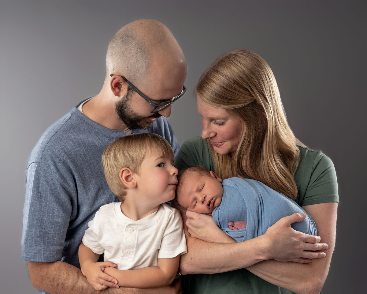 A toddler boy in a white shirt kisses his newborn baby brother with mom and dad