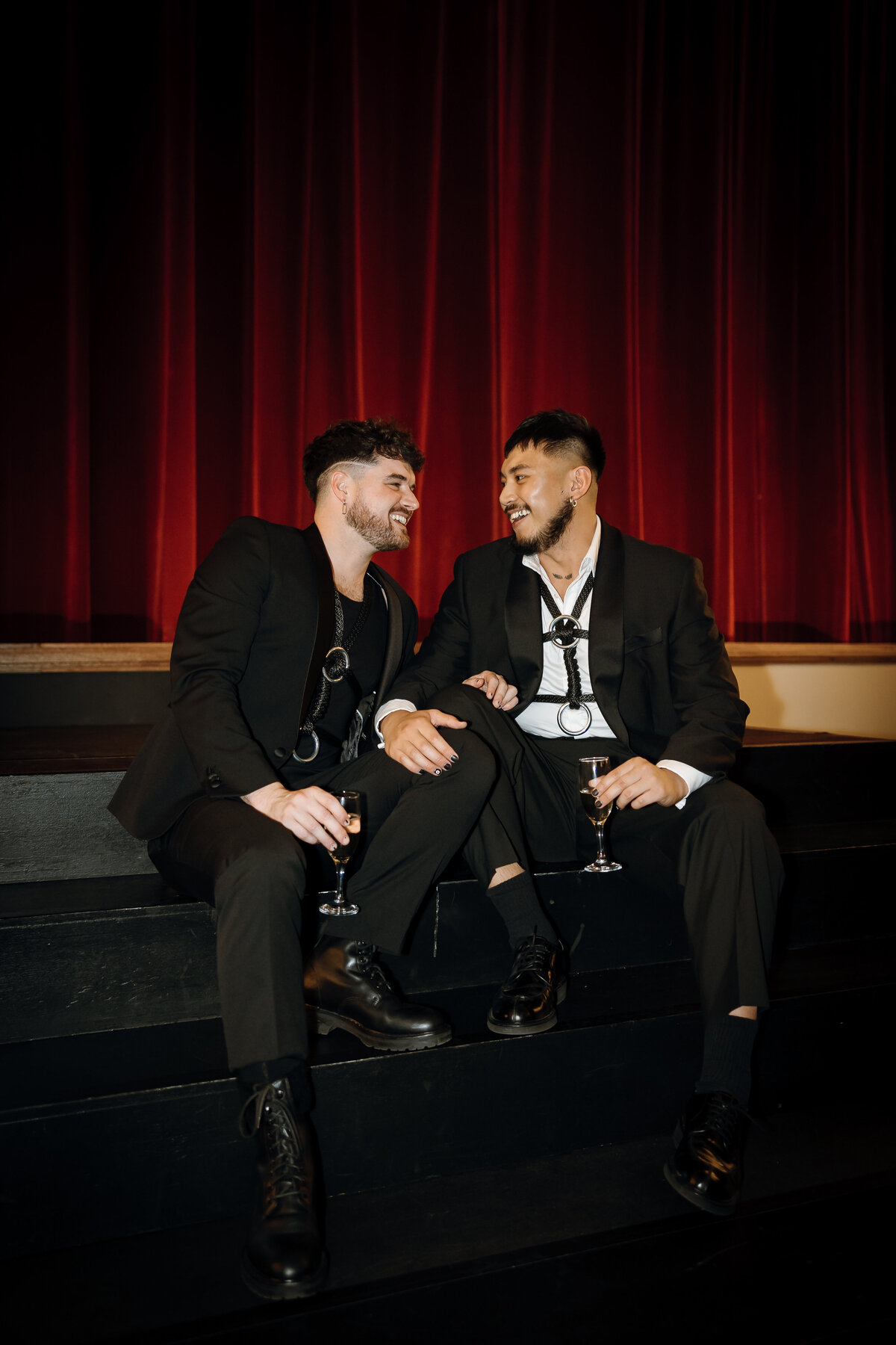 2 men in black coats smile at each other sitting on the steps of the stage.