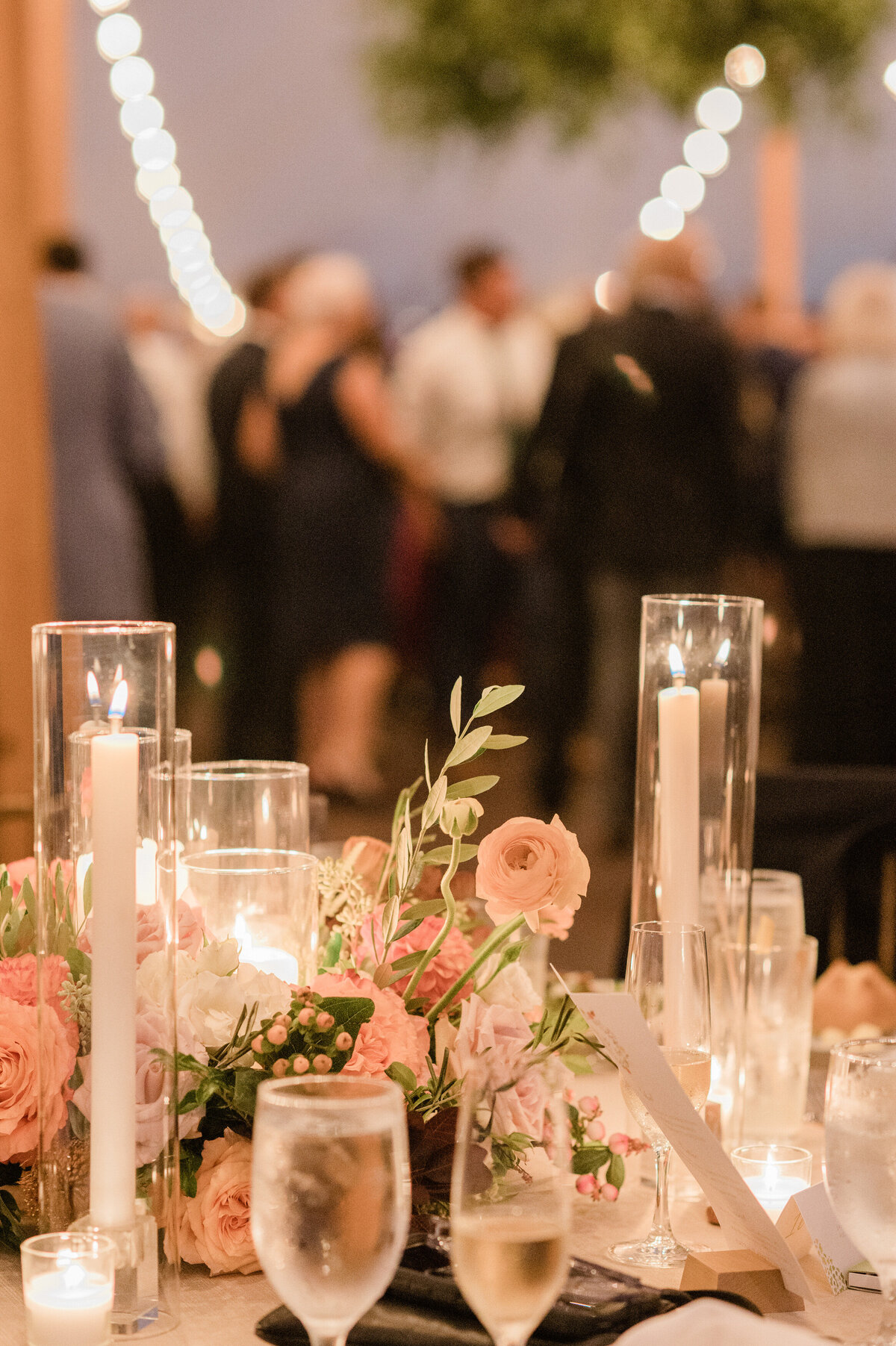 A table at Spring Lake Golf Club’s wedding reception is decorated with soft pink roses, tall candles in glass cylinders, and champagne glasses filled for toasts. Guests in formal attire mingle in the softly lit background.