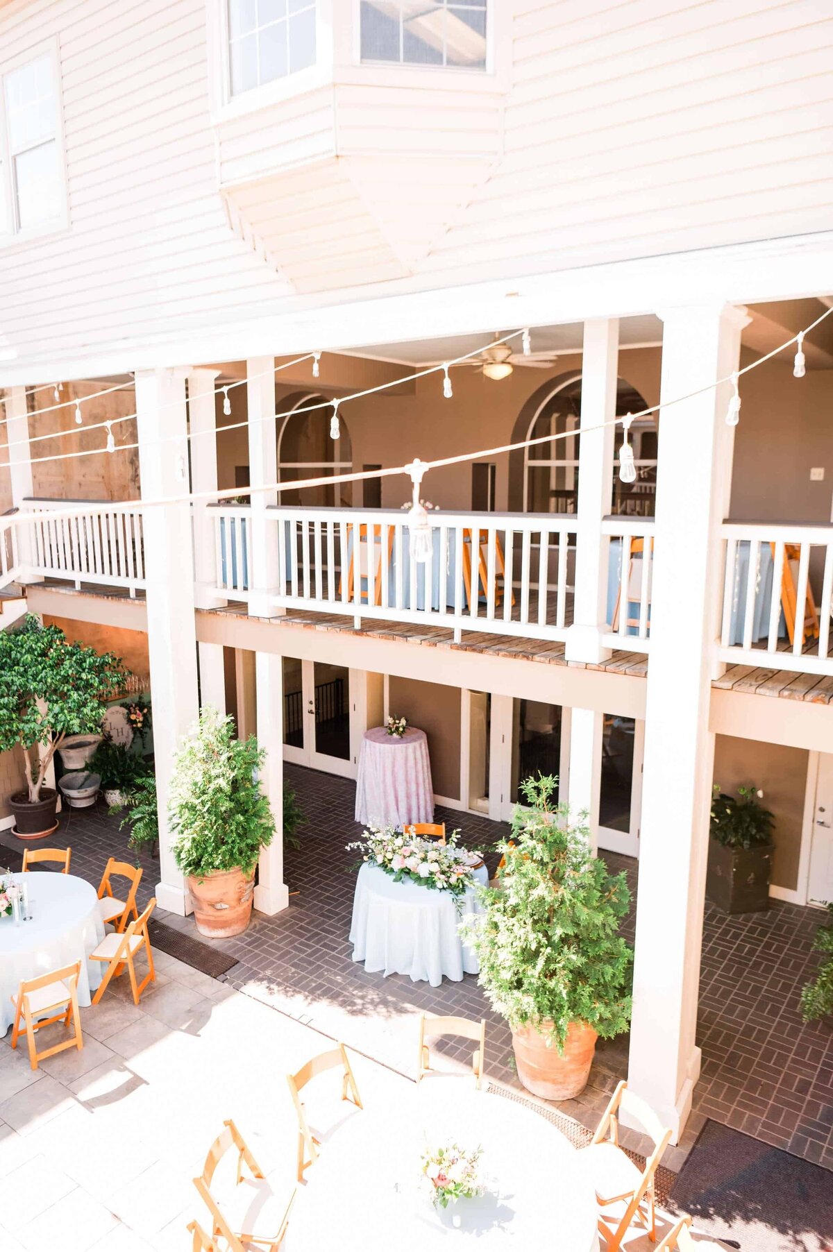 A bright courtyard set up for an event features round tables with light blue tablecloths, wooden chairs, potted plants, and string lights hanging above, bordered by two-story balconies.