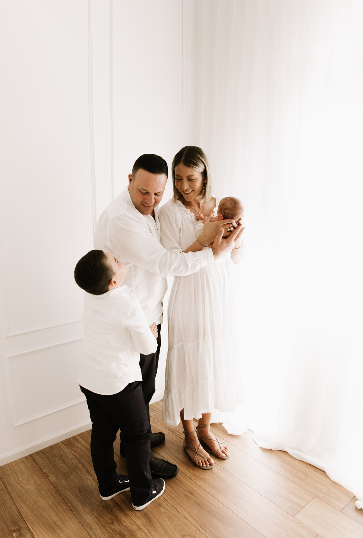 Family of four sharing a tender moment in a softly lit photography studio in Southwest Sydney during their newborn session, with both parents and siblings embracing the baby.