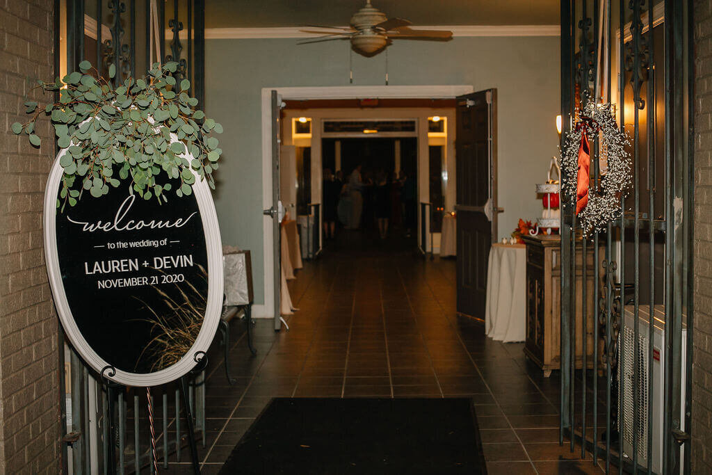 A wedding entrance with a welcome sign for Lauren and Devin. The hallway is warmly lit, decorated with greenery and autumn wreaths, leading to a room with people in the distance.