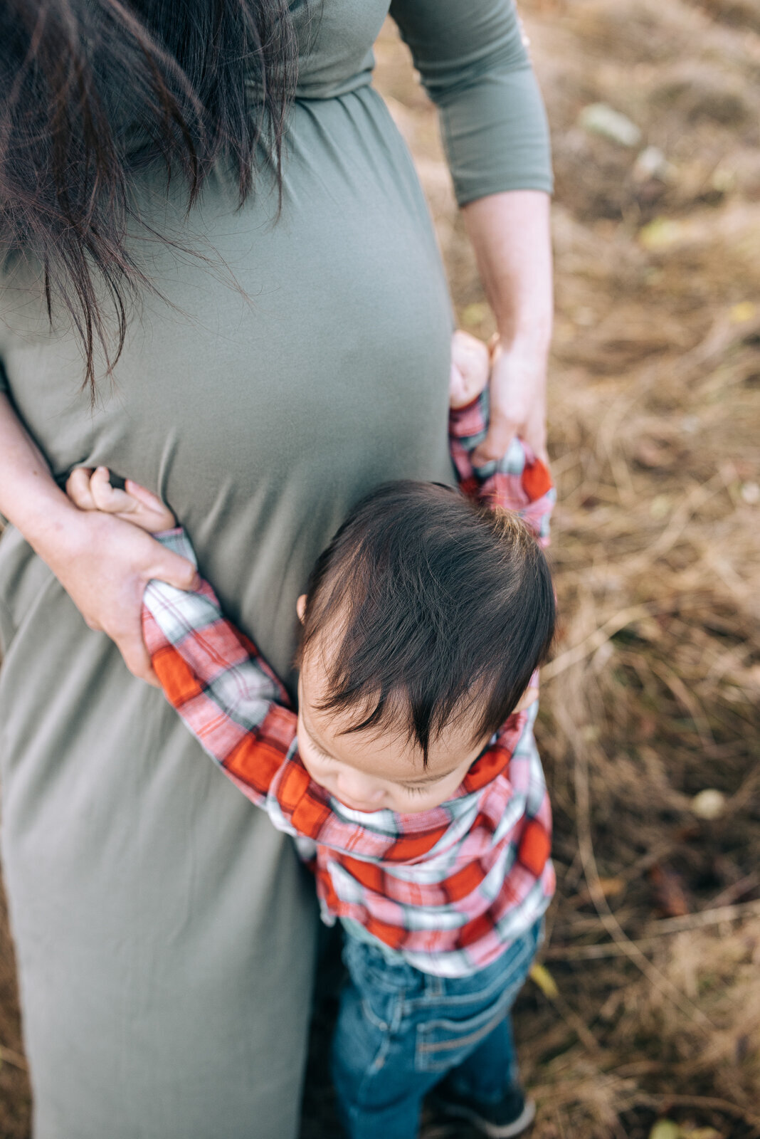 Toddler boy wearing a red plaid shirt giving his mom a hug, mom holding his hands on the side of her belly with his baby sister to be