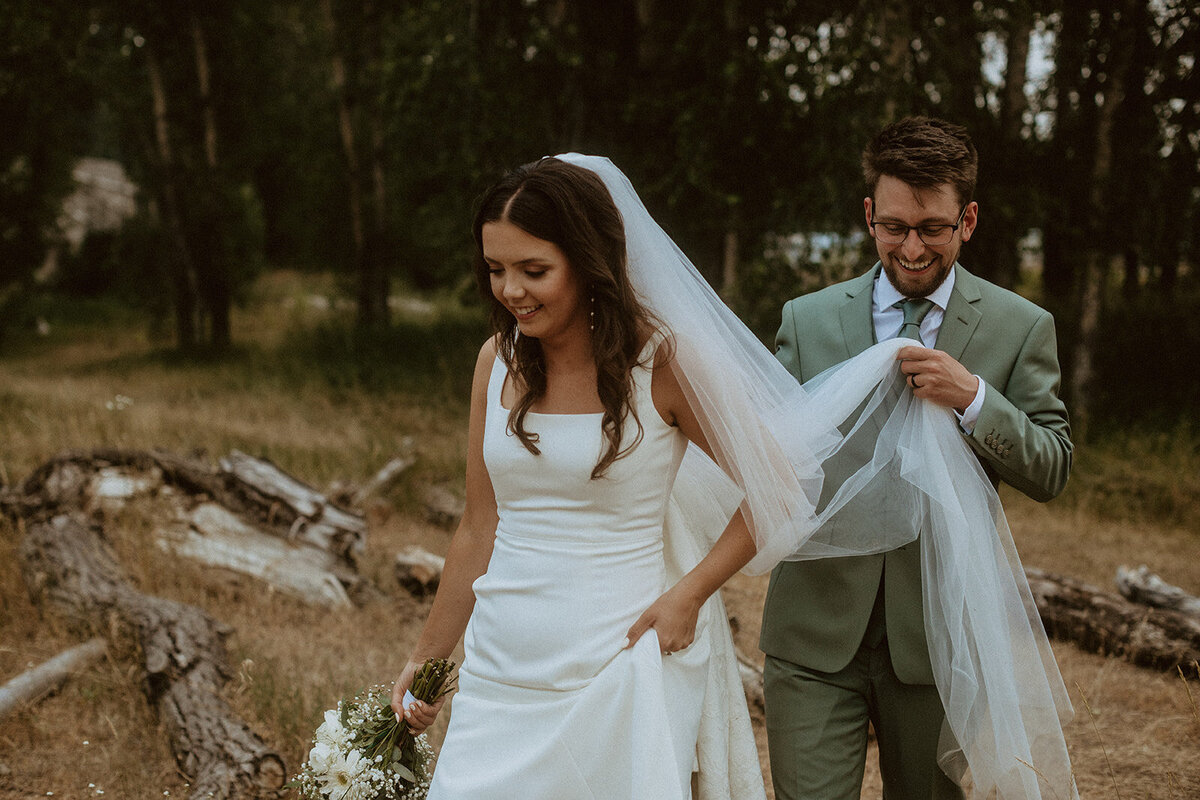 groom holds bride's veil and dress while walking
