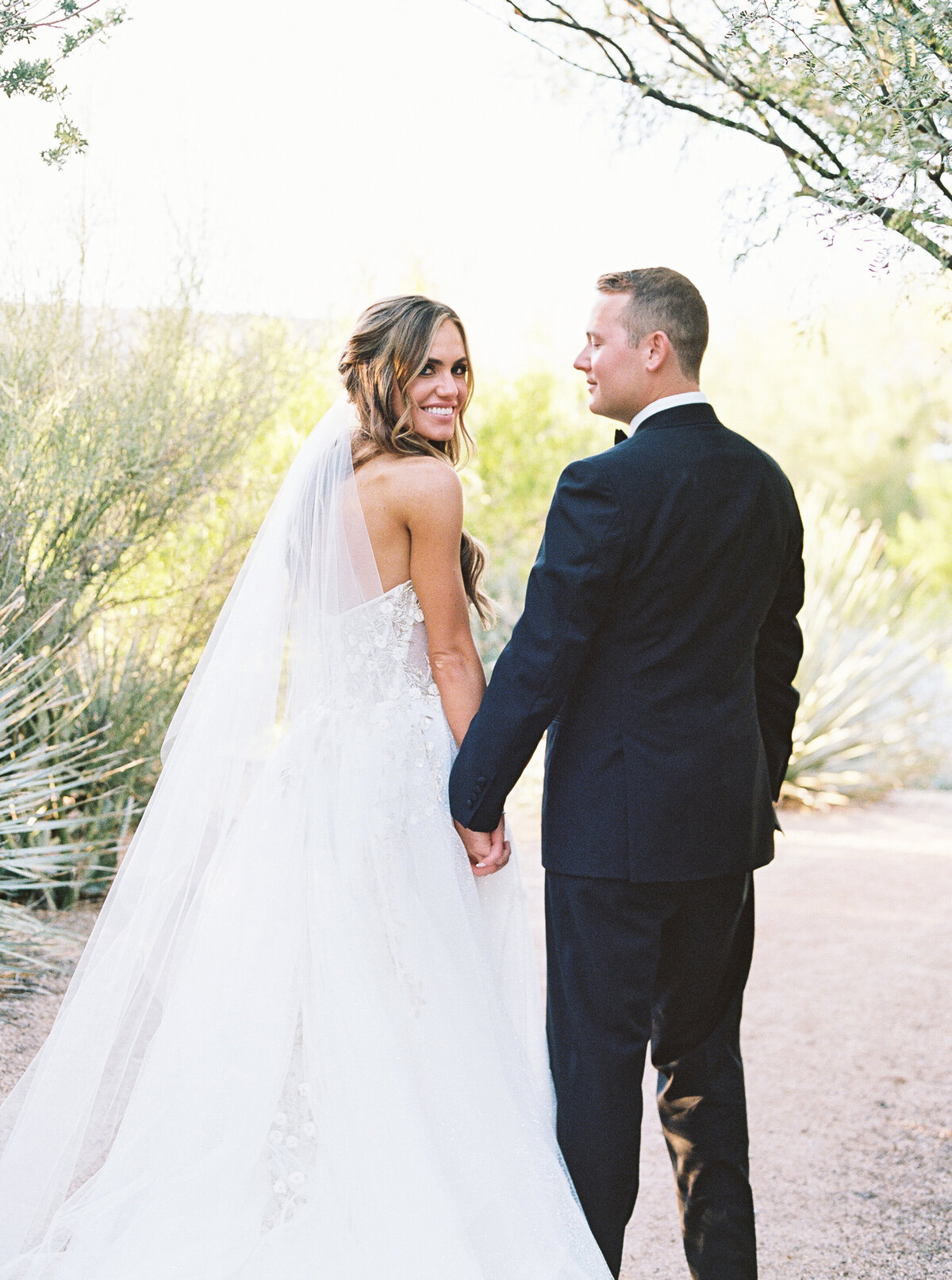 A romantic close-up of the bride and groom holding hands, showcasing the bride’s delicate veil and intricate gown details at a luxury desert wedding.