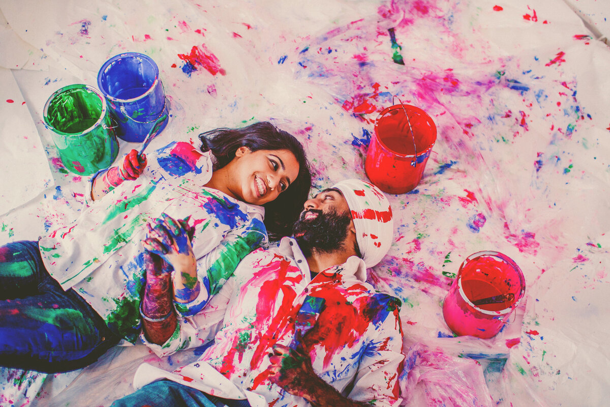 A couple laying down on a white floor with paint all over it holding hands wearing shirts with paint on it during their painting engagement session