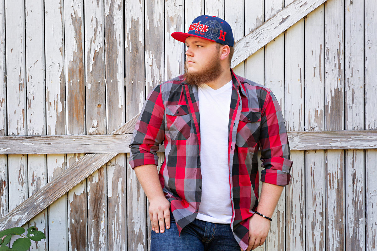 Guy in a flannel shirt and hat pose against a barn