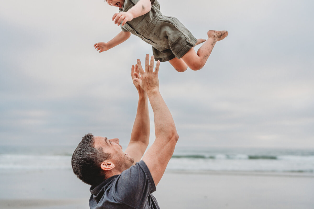 Father tossing his young daughter playfully into the air at Del Mar beach during a San Diego family photography session.