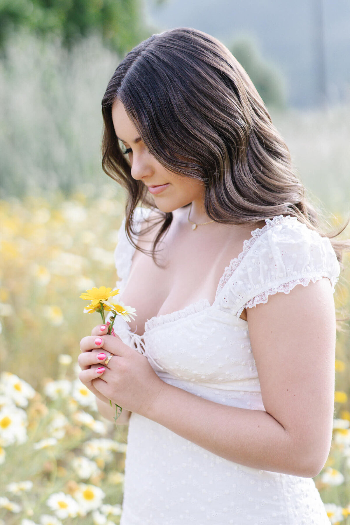 A woman with long brown hair stands in a field of daisies, holding a single yellow daisy. She is wearing a white, short-sleeved dress and gazing down at the flower, surrounded by blurred greenery in the background.