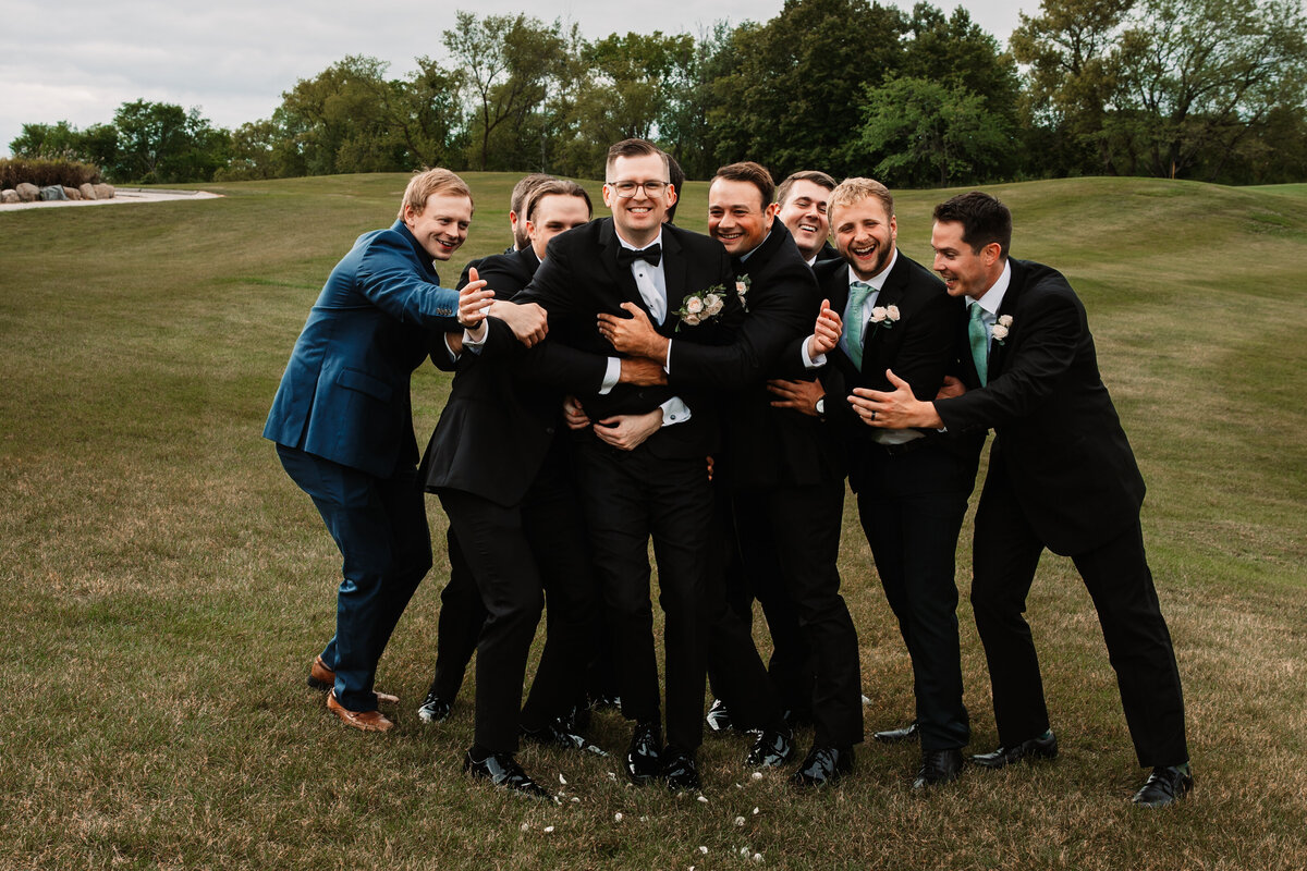 A groom is tackled by his groomsmen in a field  in black suits
