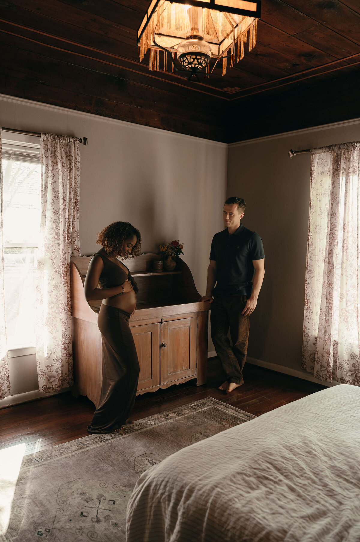 A pregnant woman tenderly touches her belly, standing beside a man in a cozy bedroom. Warm light filters through sheer curtains, creating an intimate atmosphere.