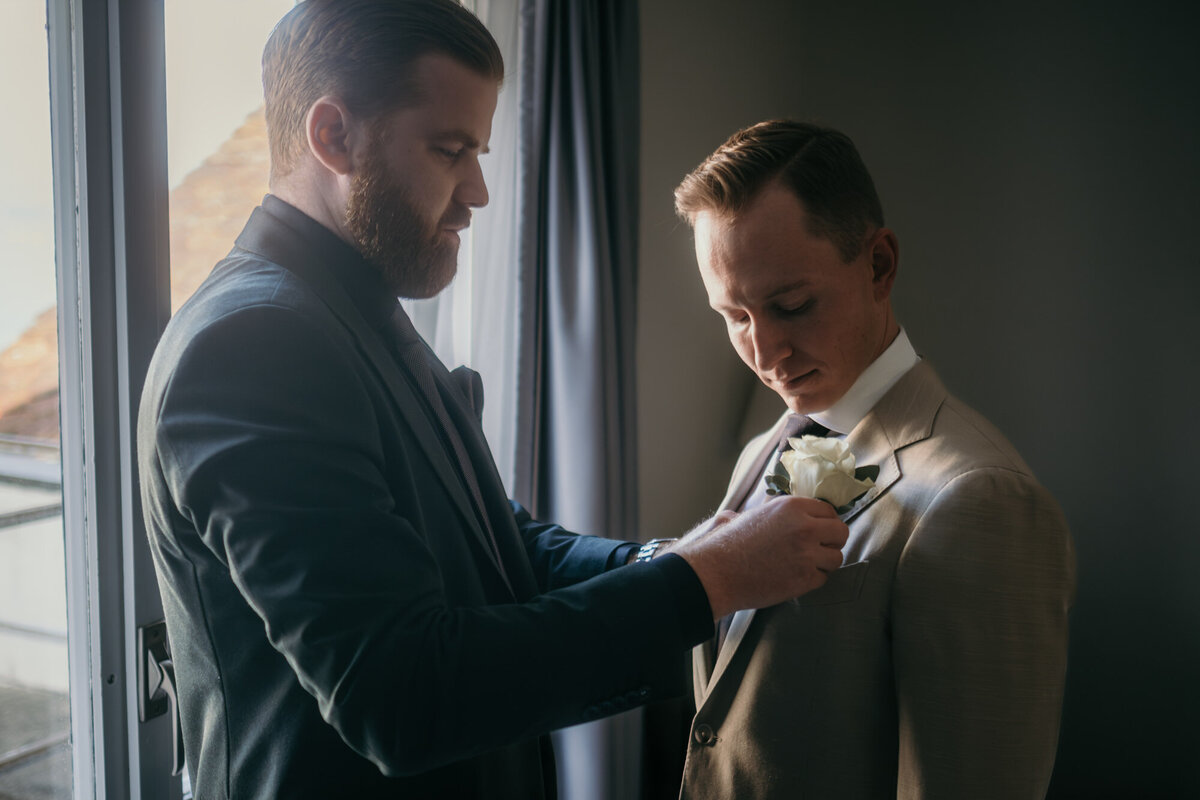 The best man adjusts the corsage flower at the grooms jacket during getting ready session.