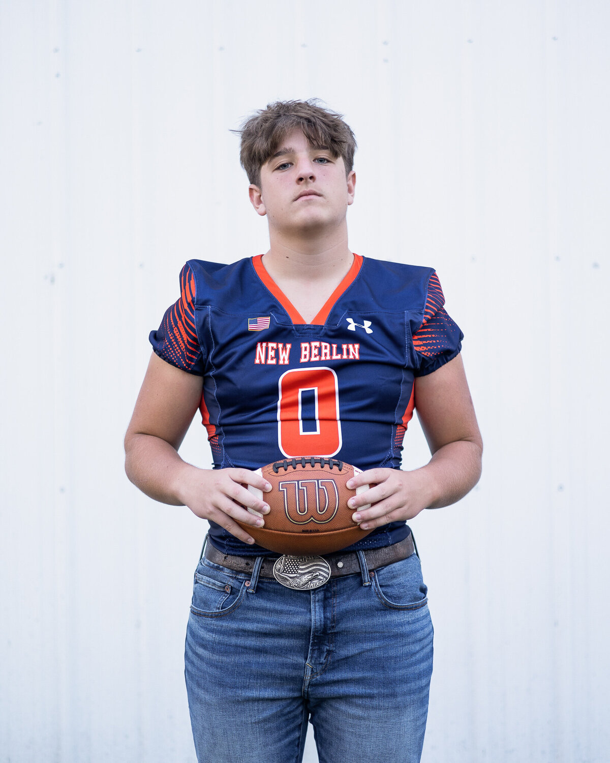 High school senior male in a navy and orange football jersey, blue jeans, belt, looking at the camera with a serious look, holding a football between his hands and has short brown hair