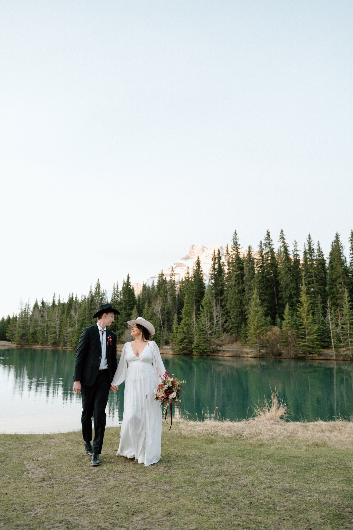 Bride and groom walking beside tranquil river with pine forest and peak reflections.