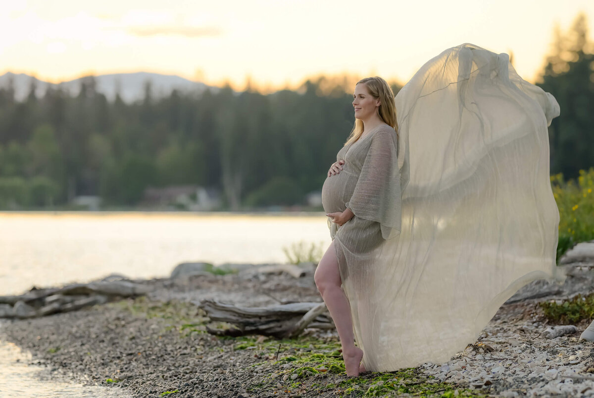 maternity photo of a mom to be PNW beach at sunset with mountains in the background. She's wearing golden sheer dress that is being gently tossed in the breeze.