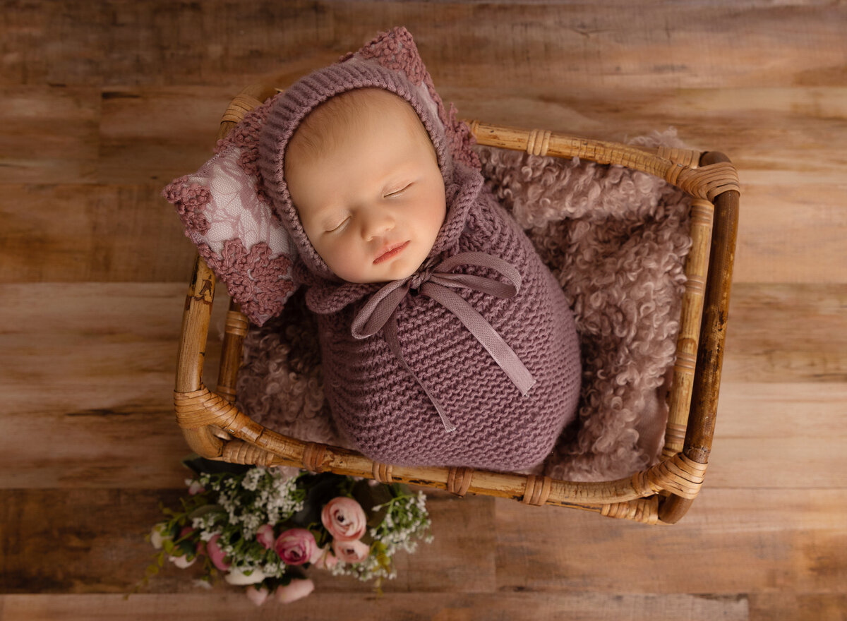 Baby girl in a mauve knit newborn posing sack is placed in a basket with a matching pillow underneath her head. there are flowers placed on the ground beside her. Captured by top Brooklyn newborn photographer Chaya Bornstein