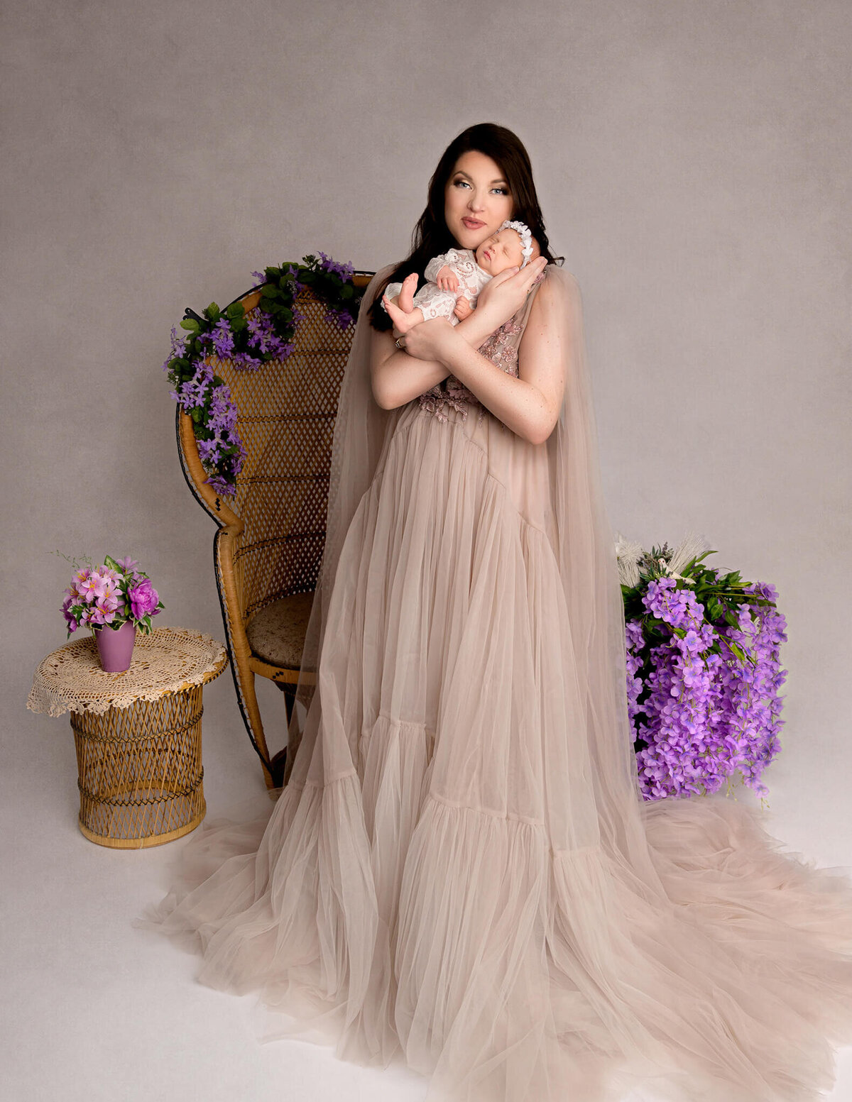 Gorgeous mom in a luxury gown holding her baby girl in her arms with boho theme and purple flowers at her photo shoot.