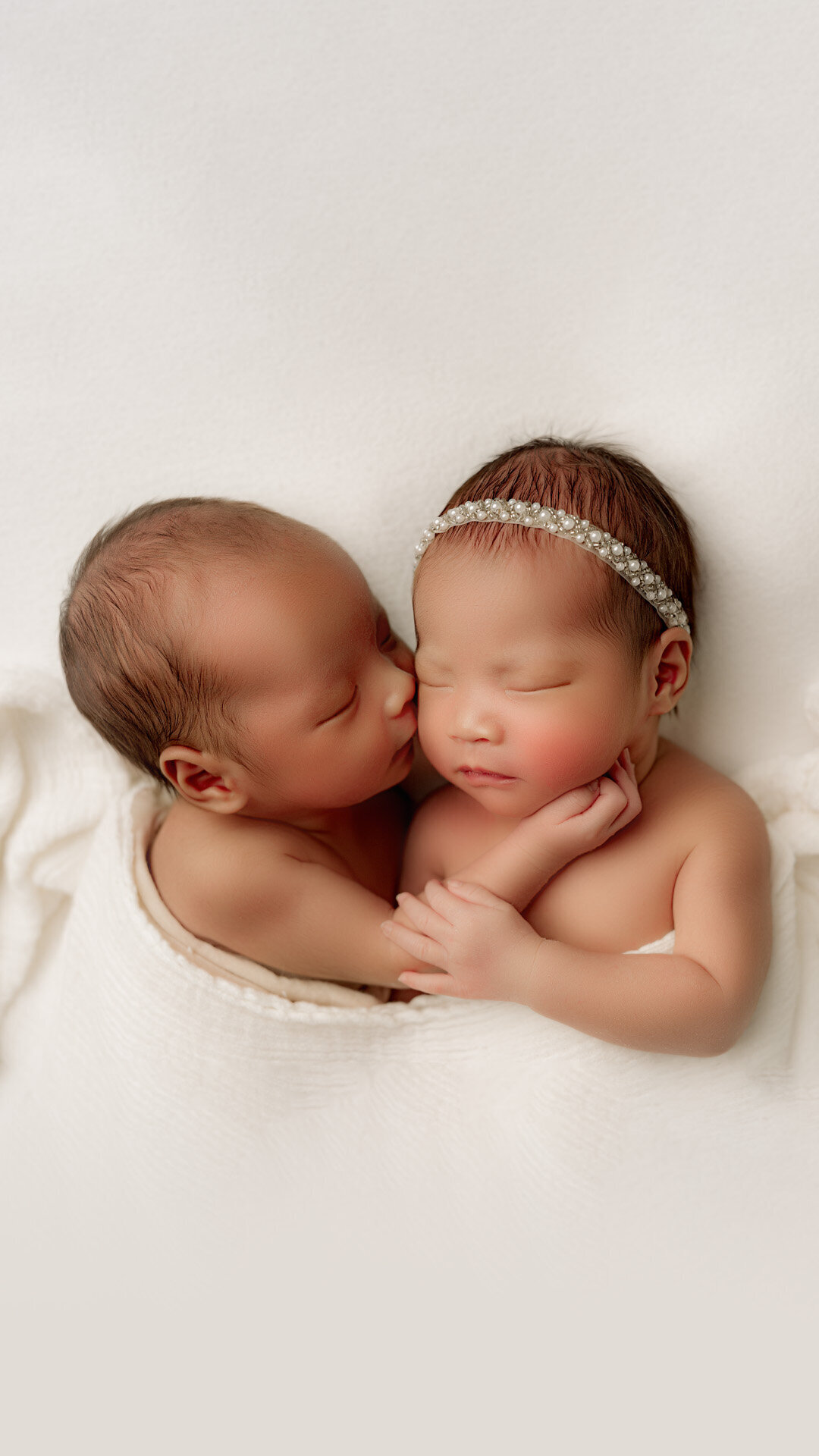 Twin newborn babies are peacefully sleeping on a soft, white blanket in a Charlotte newborn photography studio. One baby, wearing a delicate headband, is gently held by the other, who appears to be giving a sweet kiss on the cheek.