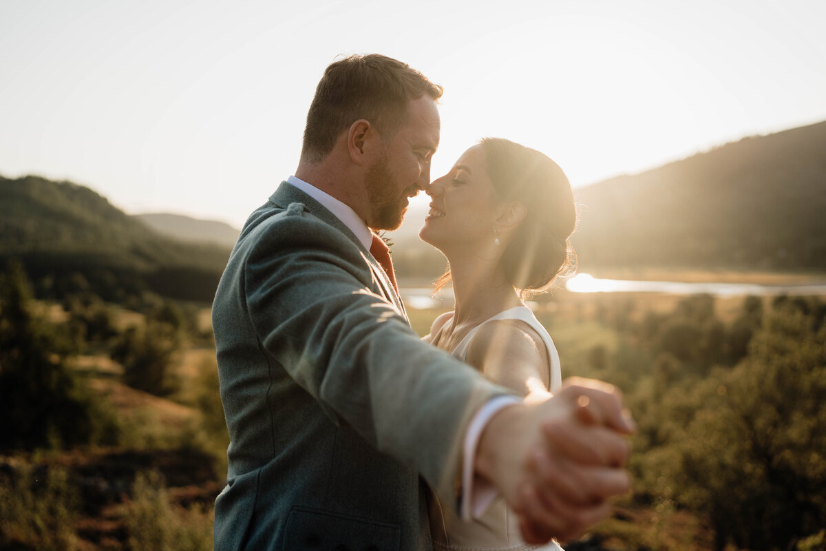 A couple link hands and press their chests together. The close photograph shows them newly married and the sun sets behind them.