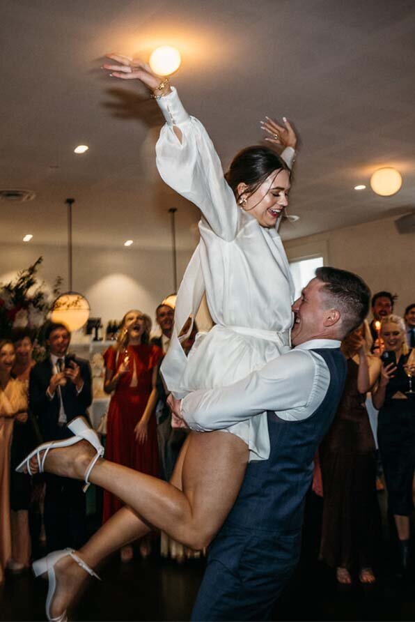 Bride smiling as the groom lifts her into the air with a warm embrace during a lively Australian wedding reception.