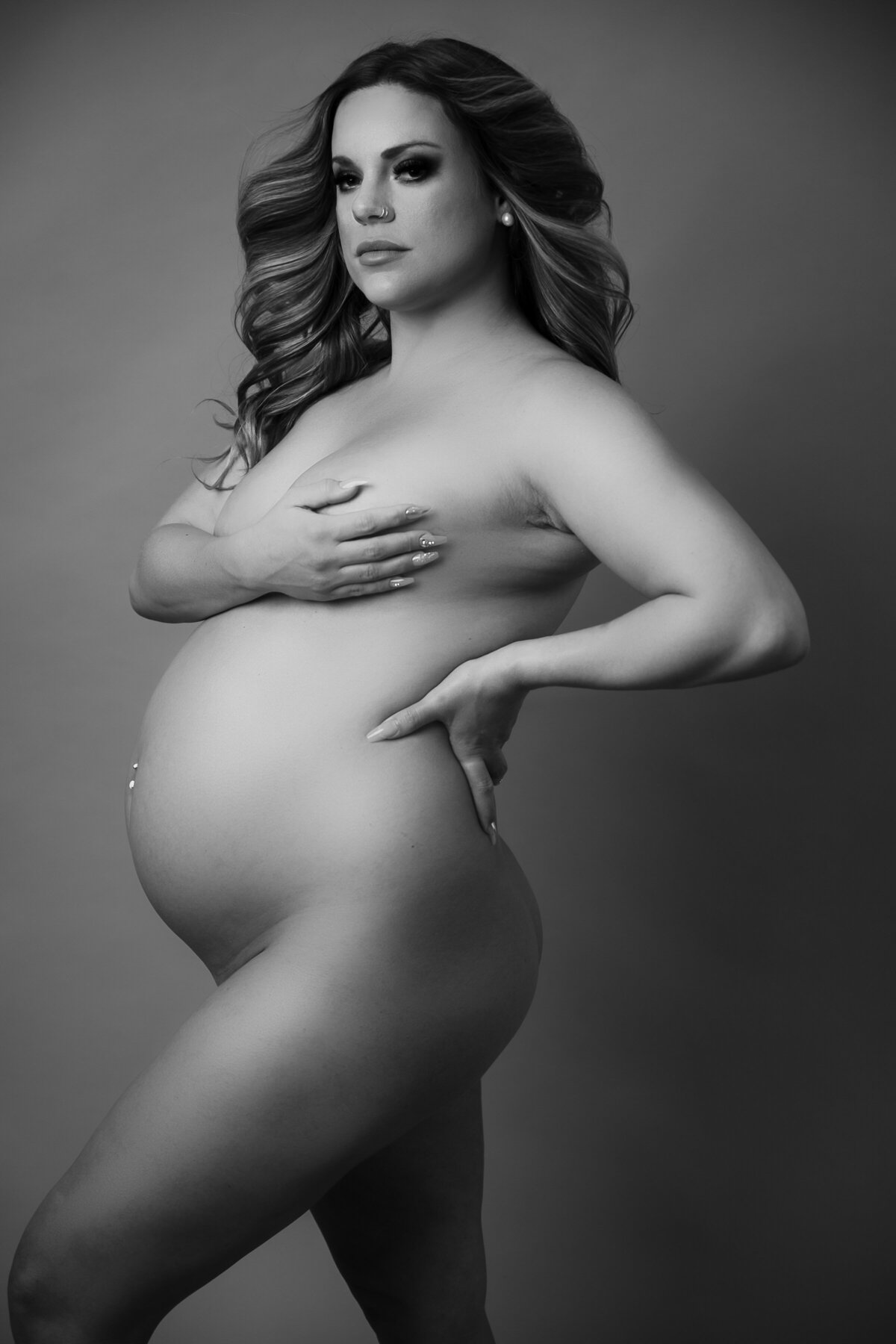 a portrait of a nude pregnant woman covering her breast with one hand while the other rest on her waist