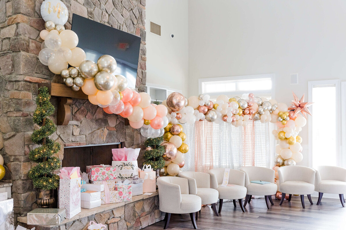 A living room, perfect for an event photographer in Fayetteville, NC, is decorated for a celebration with a large balloon garland in white, gold, and pink draped over a stone fireplace. Gift bags and wrapped presents adorn the hearth, while white chairs are arranged before sheer curtains.