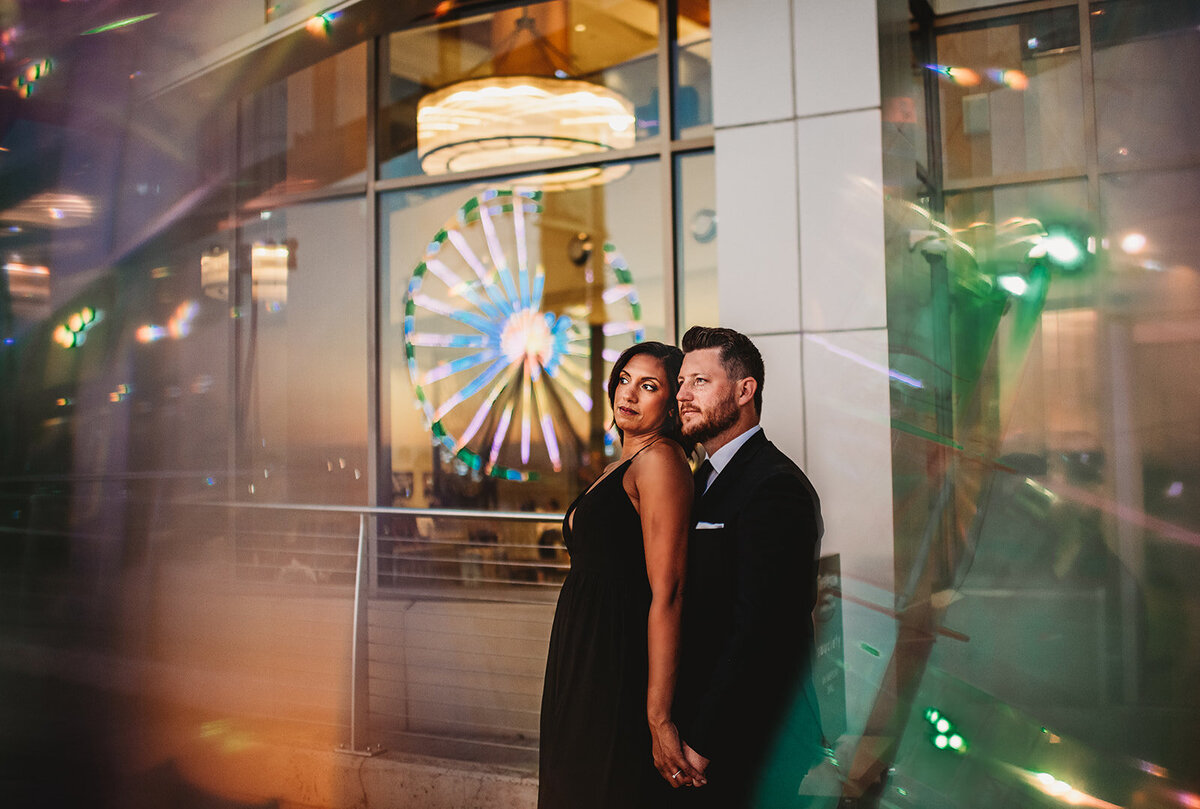 Baltimore photographers capture carnival engagement photos with woman standing in front of a man and leaning into his shoulder as they look to the bright lights of the carnival which is also reflecting in the windows behind them