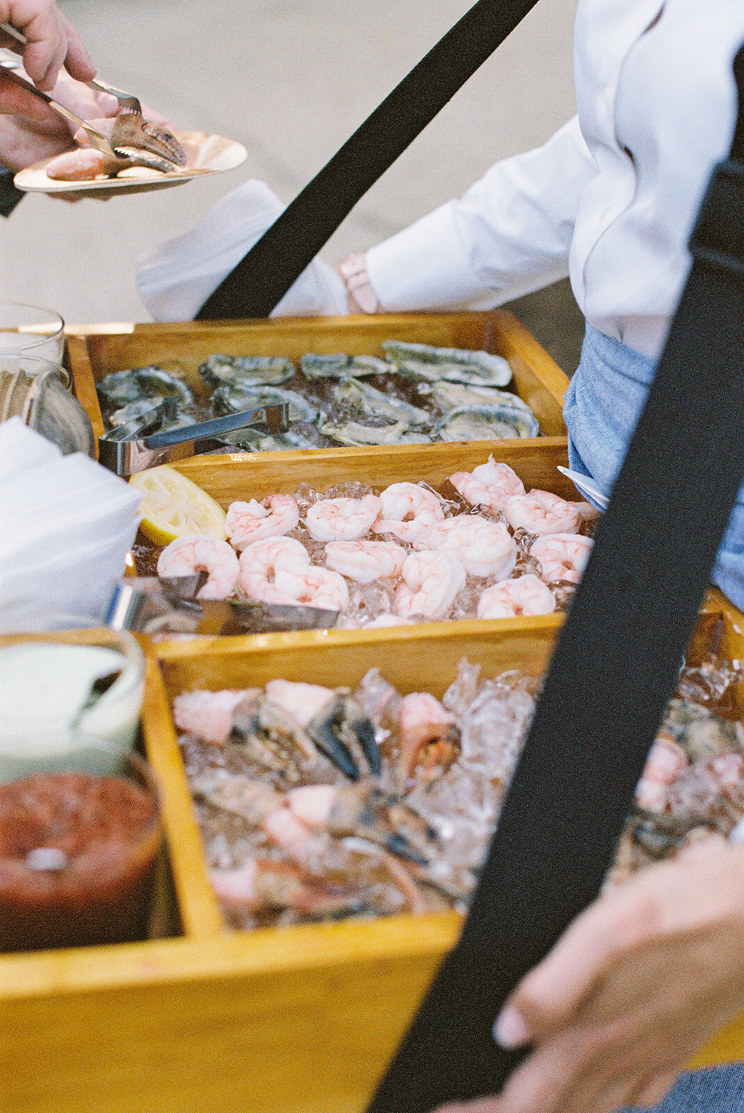 At The Hamptons, New York, waterfront estate tented wedding captured by photographer Bonnie Sen and videographers East West Films, the scene is set with a person serving a variety of fresh seafood on a wooden tray filled with ice. The seafood spread includes shrimp, mussels, and oysters, accompanied by small bowls of dipping sauces and lemon wedges. In the background, another person’s hand is seen holding a plate of delectable seafood delicacies.