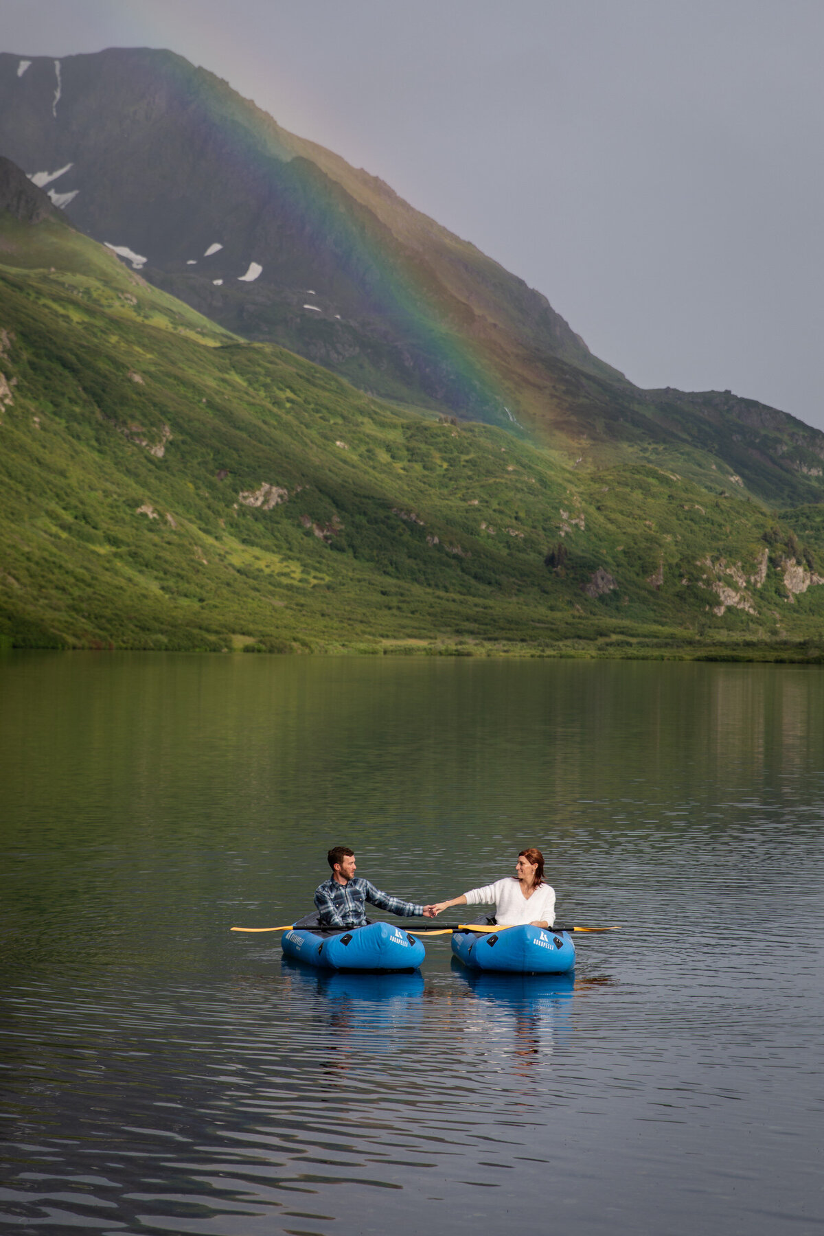 A bride and groom sit in pack rafts on a lake in Alaska holding hands
