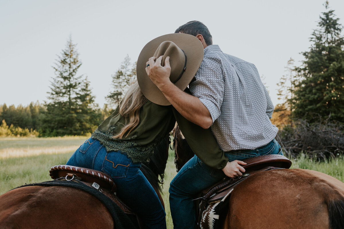 Couple on horseback lean in close and kiss behind cowboy hat.