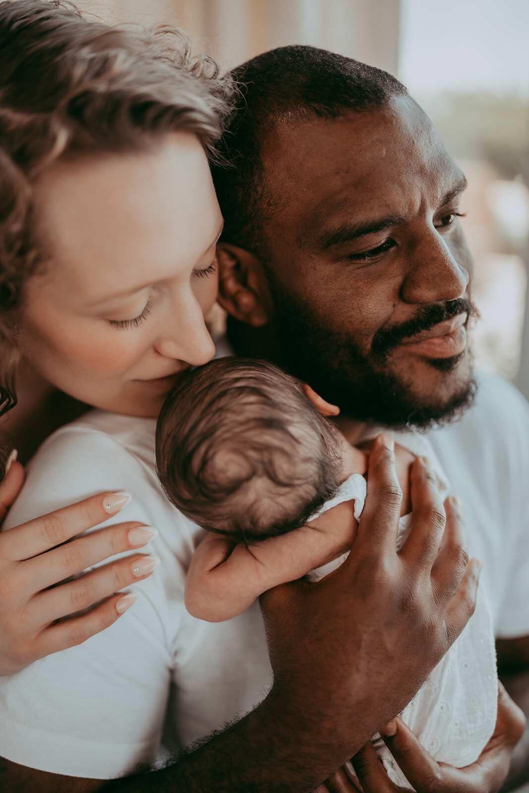 mom stands behing husband and kisses their newborn he is holding