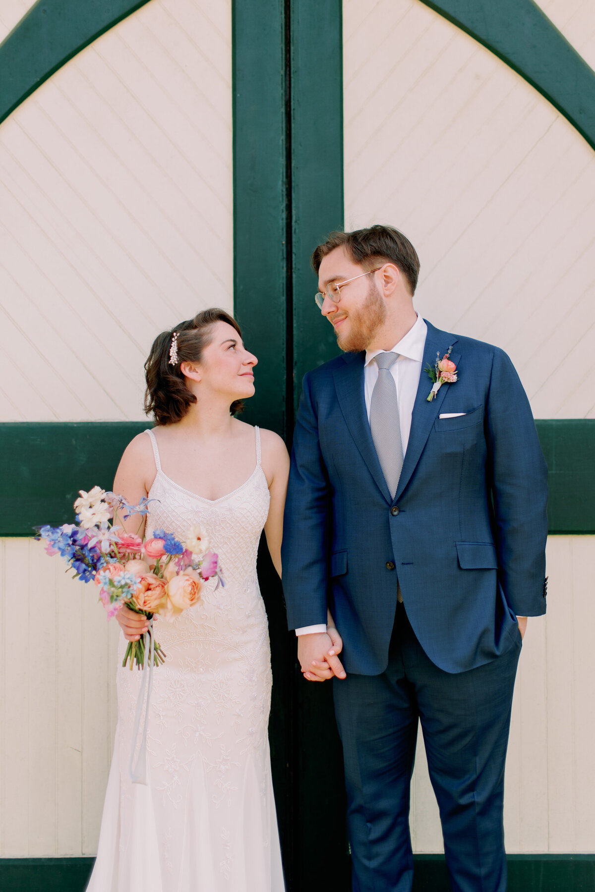 A wedding couple holding hands and standing against a wall looking at each other 