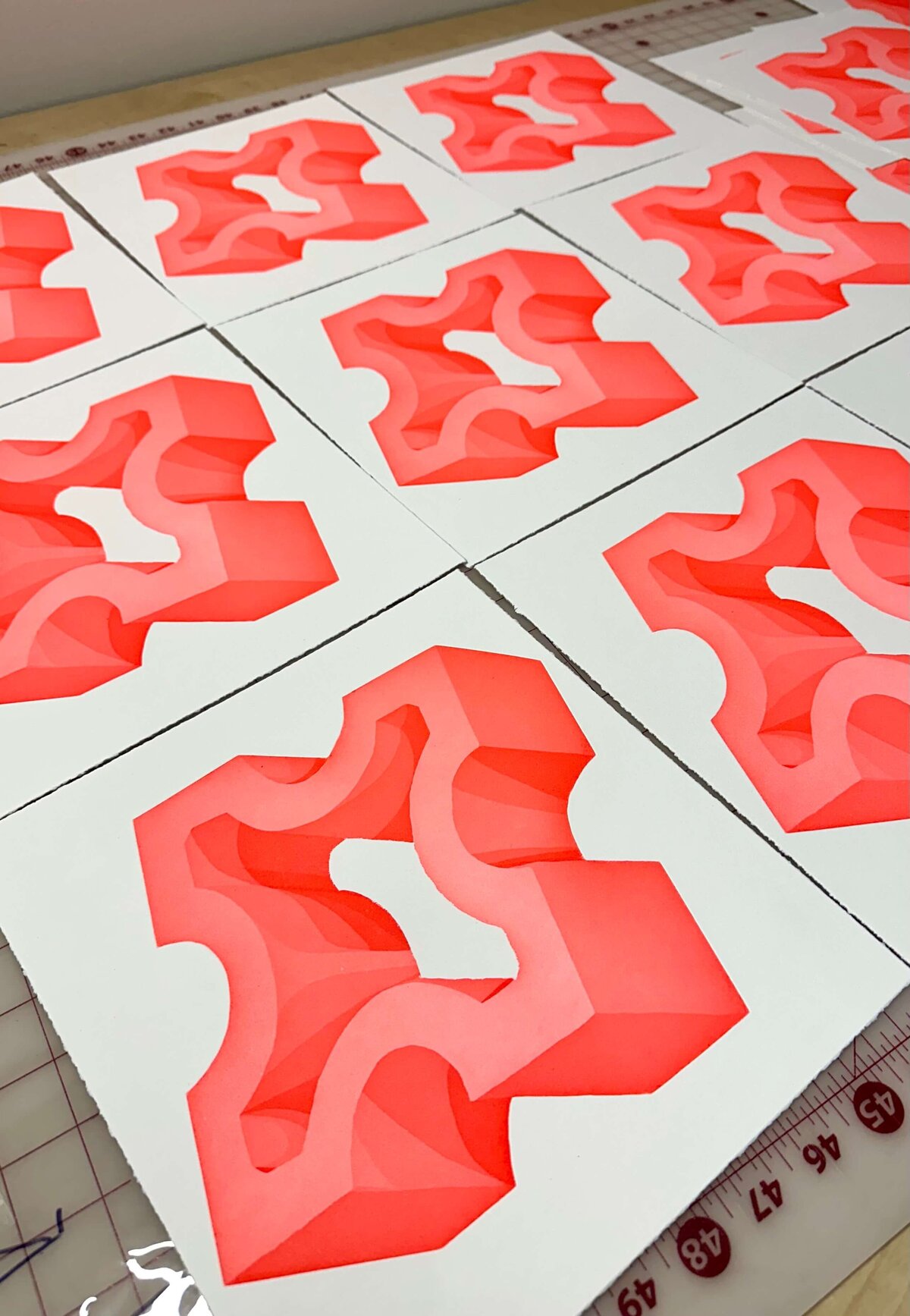Multiple red geometric drypoint prints laid out in a grid pattern on a drying rack, each showcasing dimensional shapes and smooth curves.