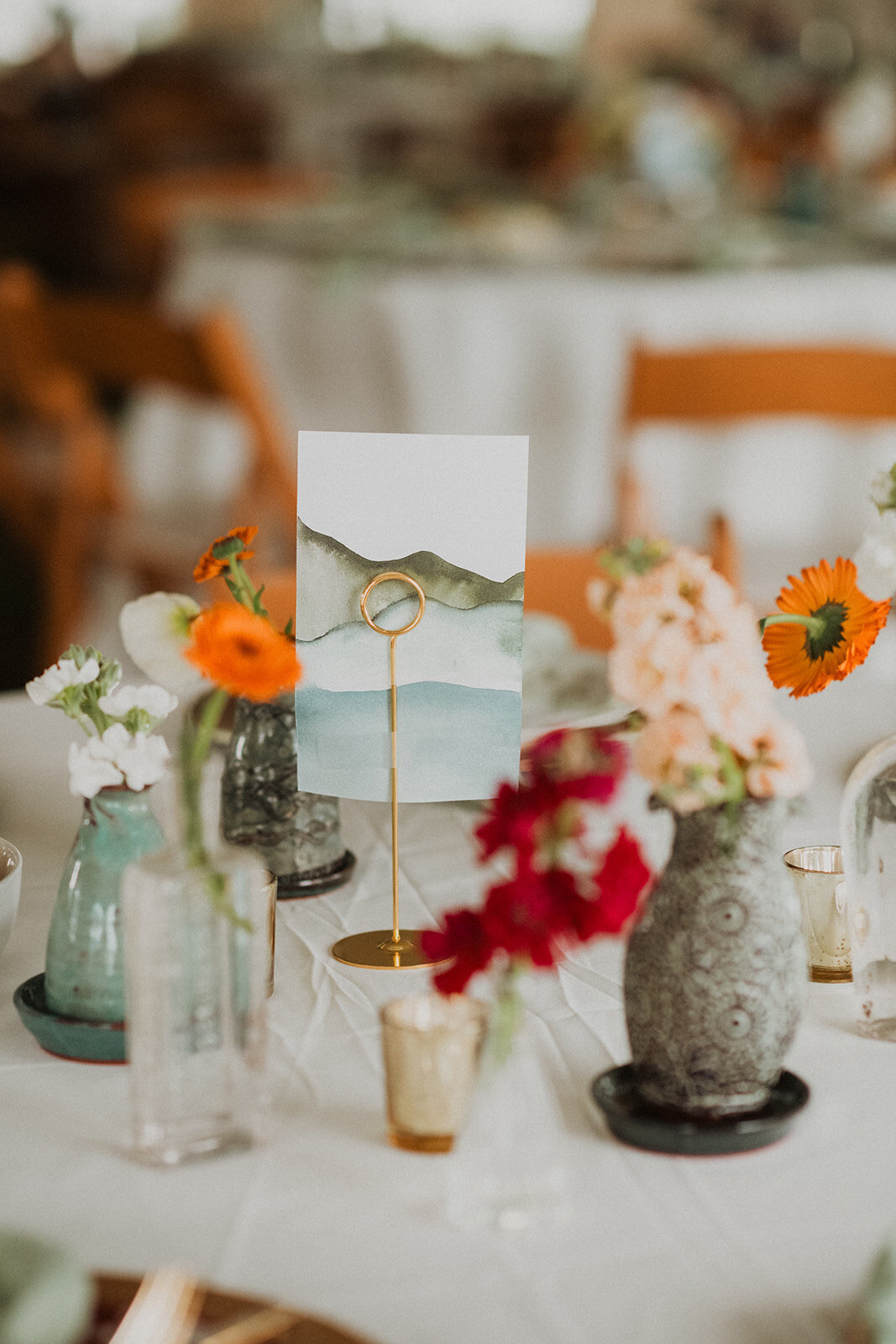 A wedding place setting with watercolor mountain cards, orange florals, and ceramic vases. 
