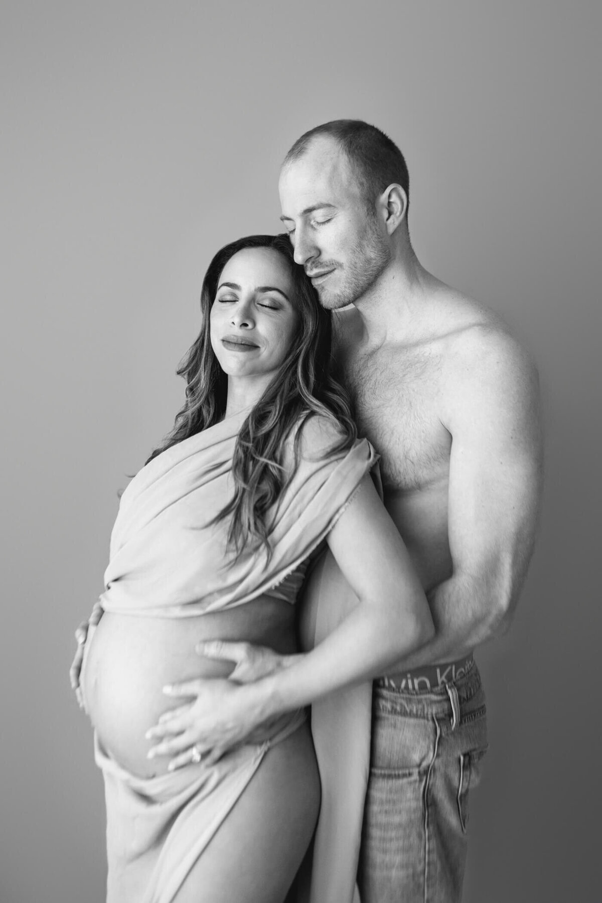 A pregnant woman stands close to her partner, both looking serene and content. The woman is wrapped in a soft, flowing fabric that accentuates her baby bump, while her partner, shirtless and wearing jeans, gently holds her from behind. Both have their eyes closed, sharing a tender moment. The black-and-white photograph emphasizes the intimacy and love between them.