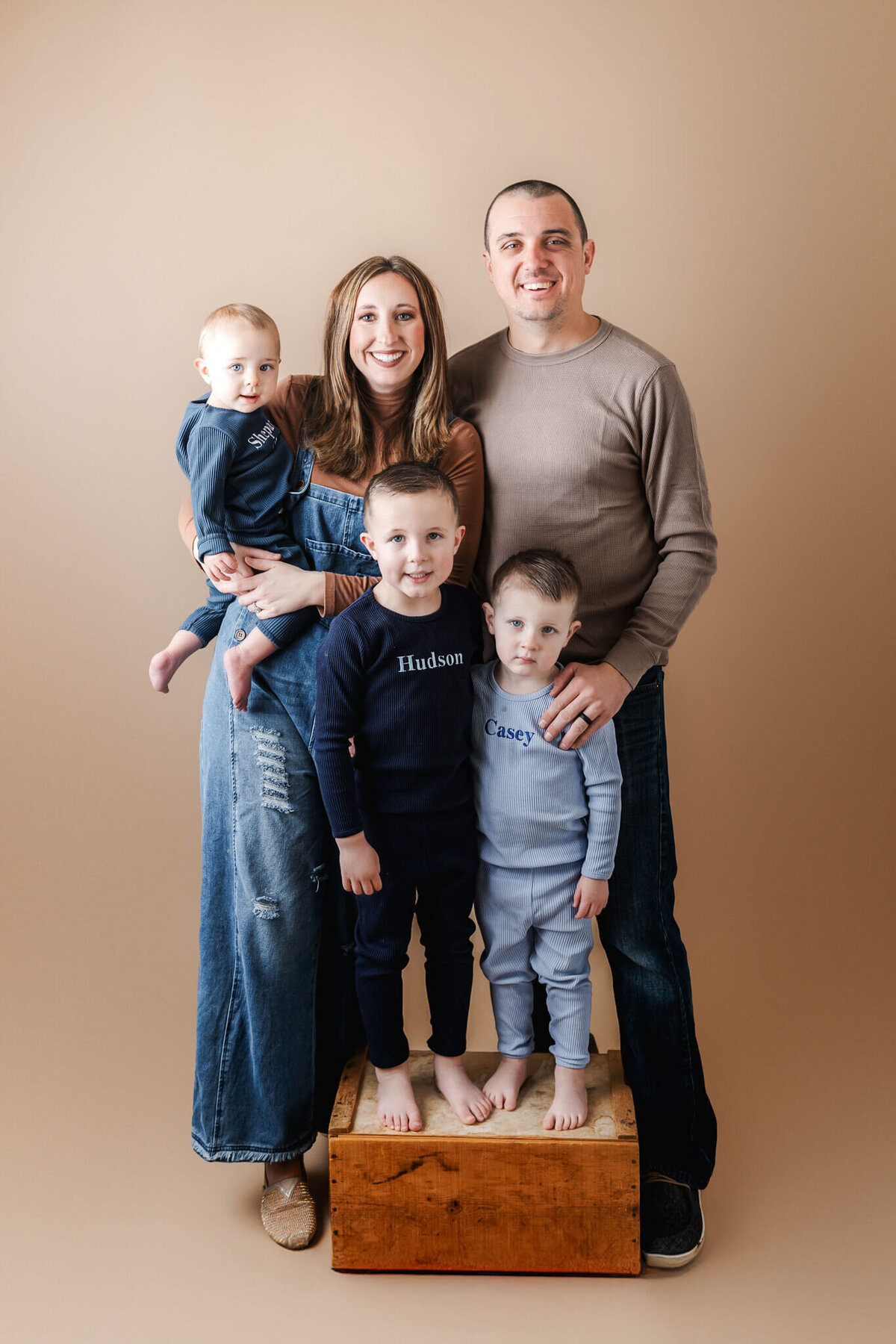 A family of five poses against a neutral background in this charming family photography session. The mother cradles a baby, flanked by two boys in personalized pajamas. With the father smiling beside them, all stand barefoot on wooden crates. Captured by a talented portrait photographer from Bloomington, Illinois.